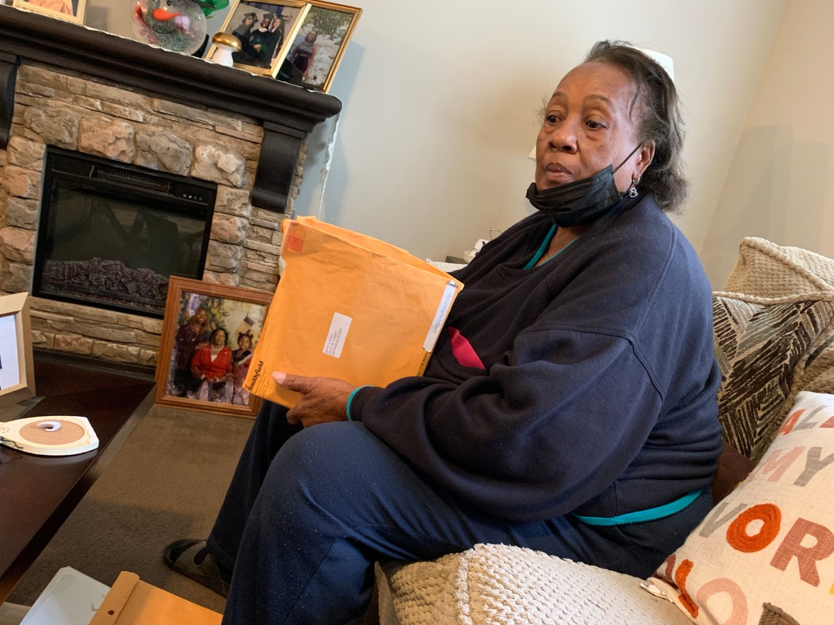 Betty Rick, a middle-aged Black woman, sits on a sofa looking distraught. She has an envelope in her hands and a mask under her chin.