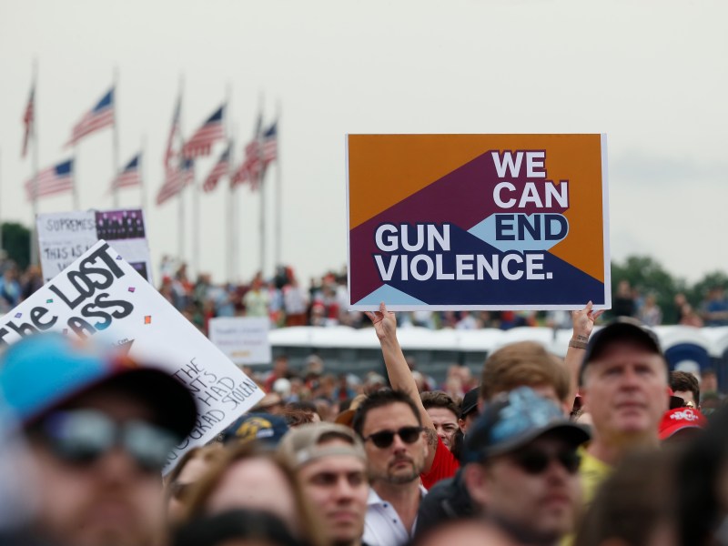 A crowd of people at a gun violence protest, their faces are out of focus. One sign that reads 'We Can End Gun Violence' is held up above the crowd.