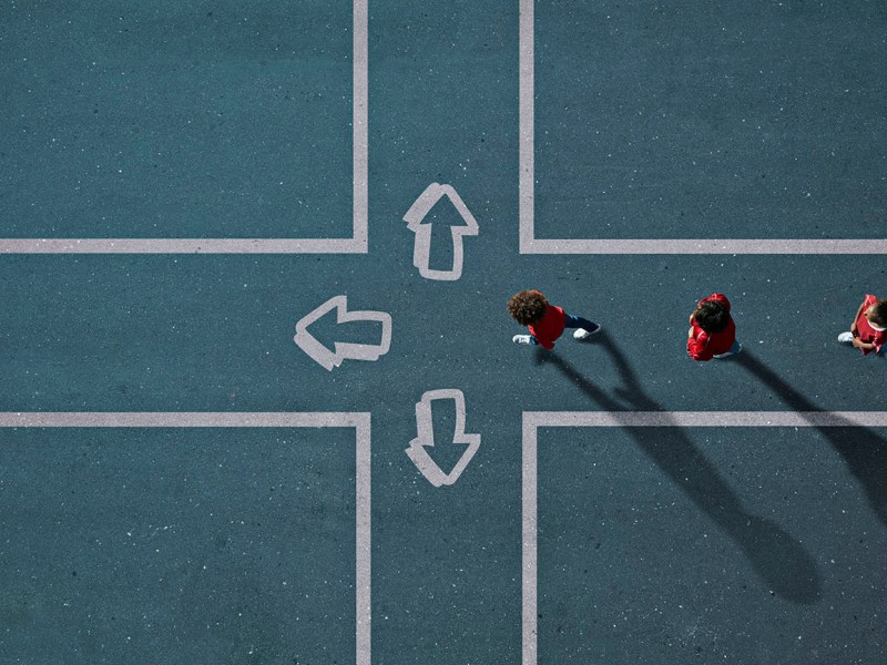 Group of children photographed from above with arrows pointing in different directions painted on the tarmac in which they walk.