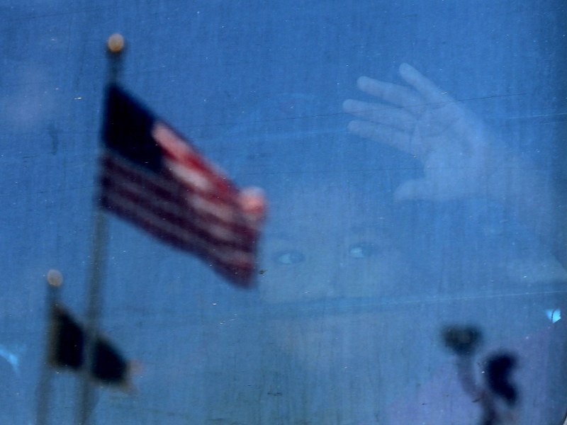 A child looks out of the window of a bus, in the reflection of the window you can see two American flags.