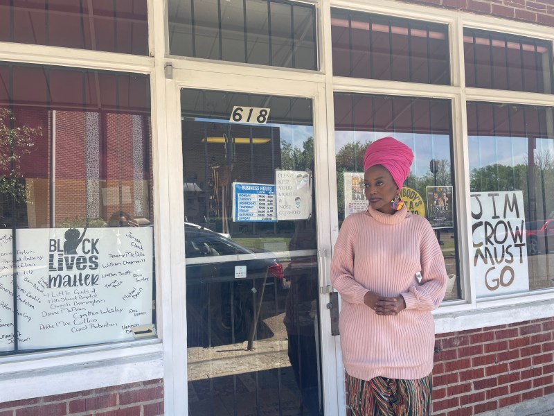 Maati Jone Primm stands in front of her store. She is wearing a pink outfit, and she has two signs in the windows of her store. One says "Jim Crow Must Go" and the other says "Black Lives Matter."