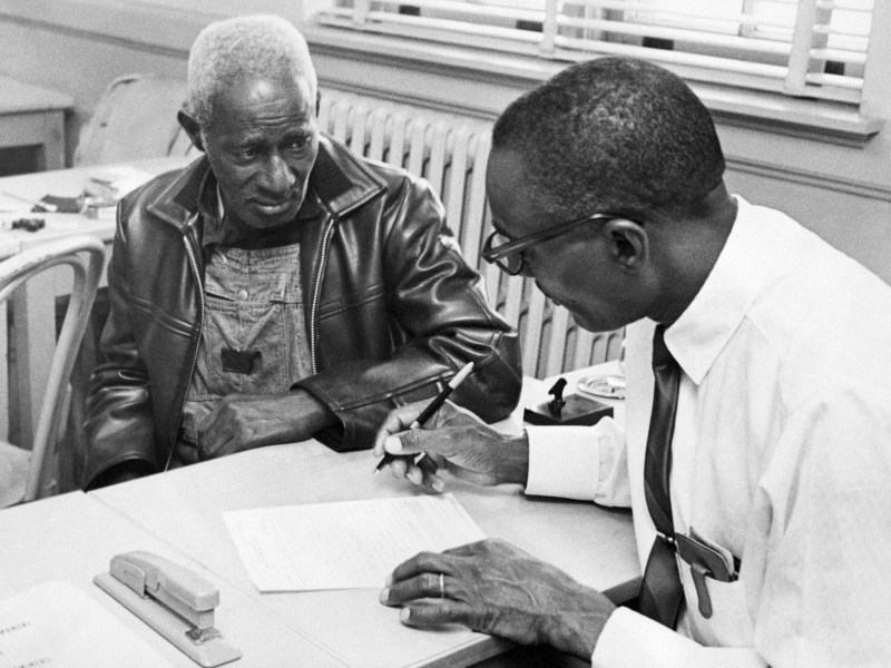 Two Black men sit at a table, going over paperwork together. There's a window in the background.