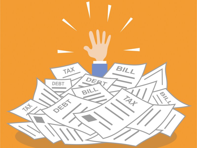 A person's hand is reaching out of an overwhelming pile of papers that say "tax," "debt" and "bill."