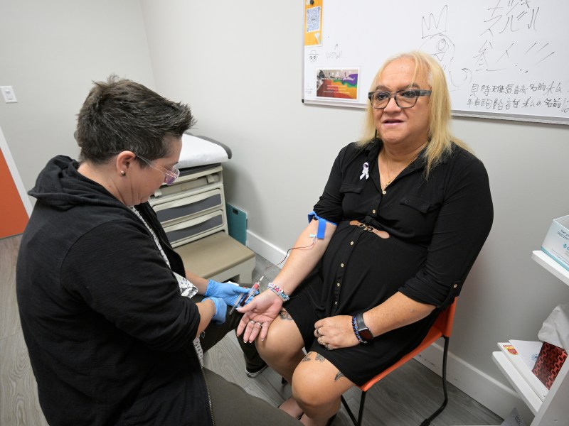 A medical personnel, dressed in a black hoodie, draws blood from Andrea Montanez, dressed in a black dress, as she sits a medical office.